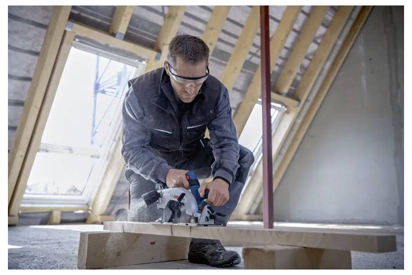 Eine Person mit Schutzbrille verwendet eine Kreissäge, um ein Holzbrett in einem teilweise fertiggestellten Gebäude zu schneiden.