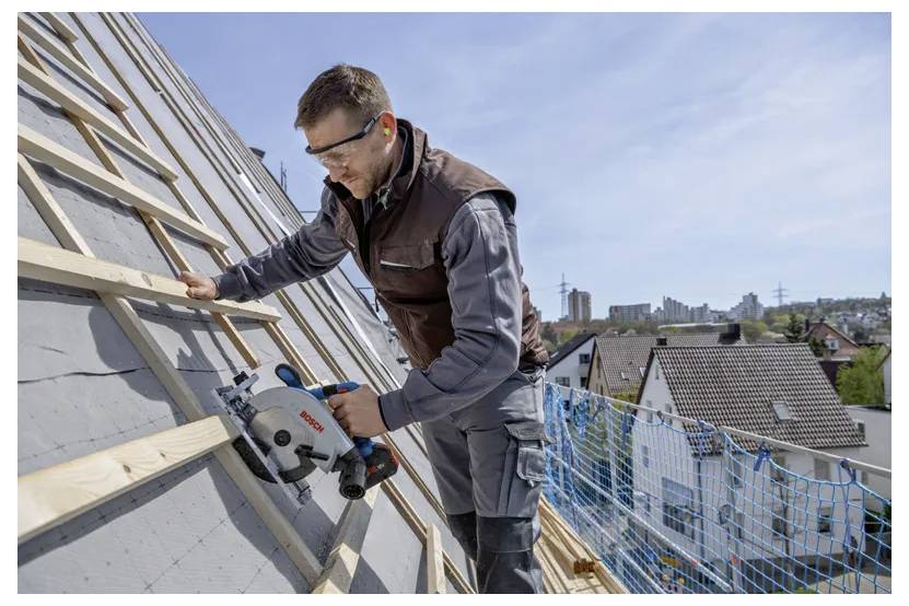 Arbeiter auf einem geneigten Dach, der eine Kreissäge verwendet, um einen Holzbalken zu schneiden, trägt Sicherheitsbrille und Warnweste, mit Stadtgebäuden im Hintergrund.