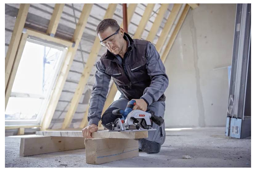 Eine Person mit Schutzbrille schneidet Holzbretter mit einer elektrischen Säge in einem unausgebauten Dachboden, umgeben von freiliegenden Balken.