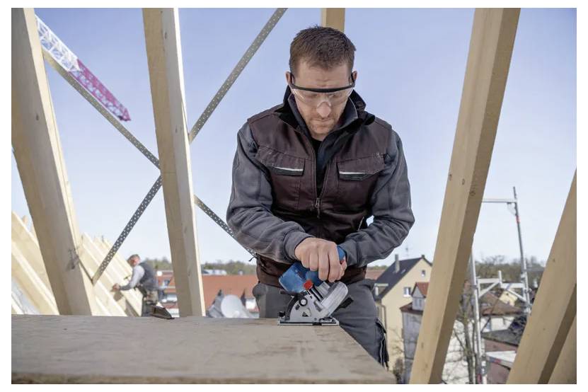 Ein Bauarbeiter mit Schutzbrille bedient ein Elektrowerkzeug an einem Holzbalken auf einer Baustelle, wobei Dächer im Hintergrund zu sehen sind.