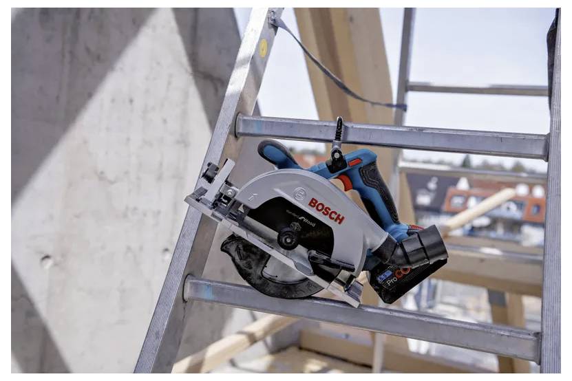 Eine Bosch-Kreissäge ist an einer Metallleiter auf einer Baustelle montiert. Im Hintergrund sind Holzbalken und ein Gebäude zu sehen.