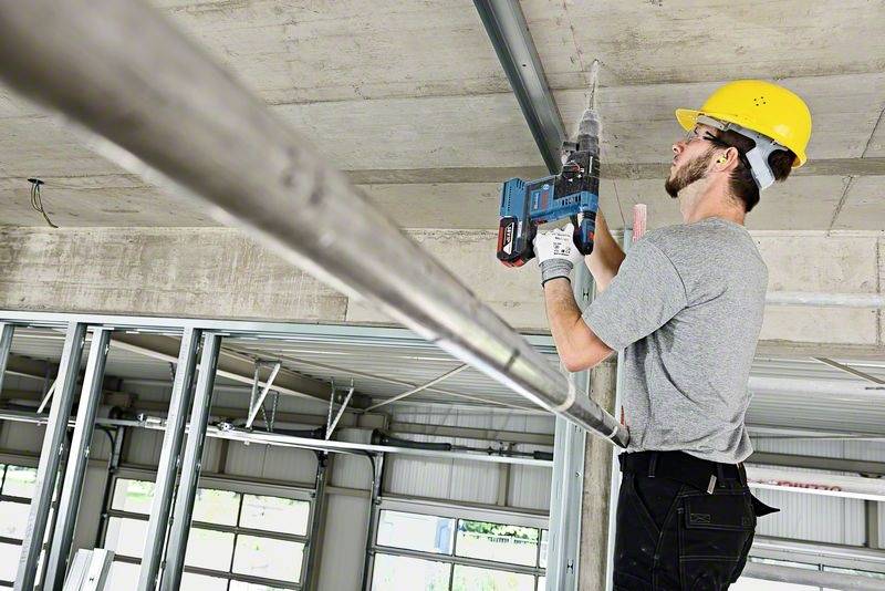 Arbeiter mit Schutzhelm und Bohrmaschine befestigt Metallrahmen an Decke in einem Rohbaugebäude.
