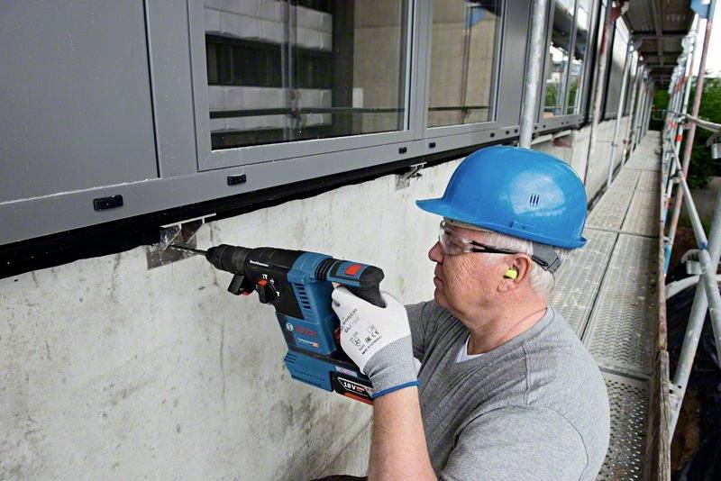 Ein Arbeiter in Schutzhelm und Handschuhen befestigt eine Metallkonstruktion an einer Betonwand mit einem elektrischen Bohrhammer.