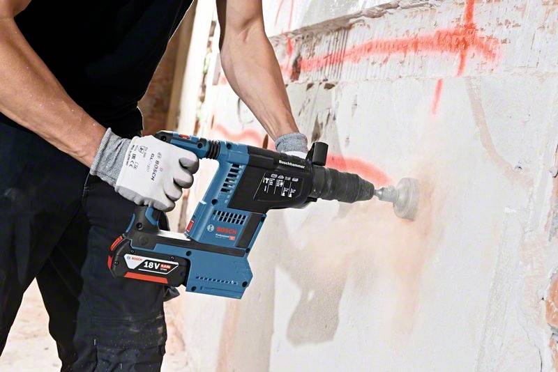 Person arbeitet mit einem elektrischen Bohrhammer, bohrt ein Loch in eine Betonwand, trägt Schutzbekleidung. Baustellenumgebung.