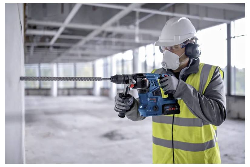 Arbeiter in Schutzausrüstung verwendet eine Bohrmaschine an einer Betonwand in einem großen, unfertig wirkenden Gebäude.