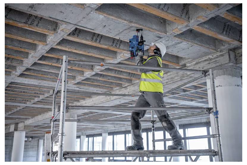 Ein Bauarbeiter in einer Sicherheitsweste und einem Helm bedient ein Elektrowerkzeug an einer Decke, während er auf Gerüsten in einem unfertig gebauten Gebäude steht.