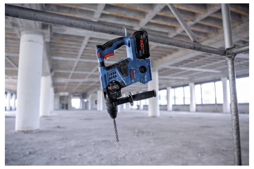 Ein blauer schnurloser Bohrhammer ist an einem Metallträger in einem großen, unfertig wirkenden Baustellenraum mit Betonsäulen und Betonböden befestigt.