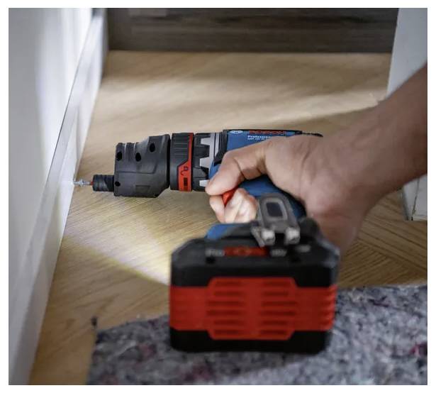 Eine Hand hält einen kabellosen Akkuschrauber und schraubt in einen Sockelleiste auf einem Holzboden, wobei ein grauer Teppich teilweise im Vordergrund sichtbar ist.