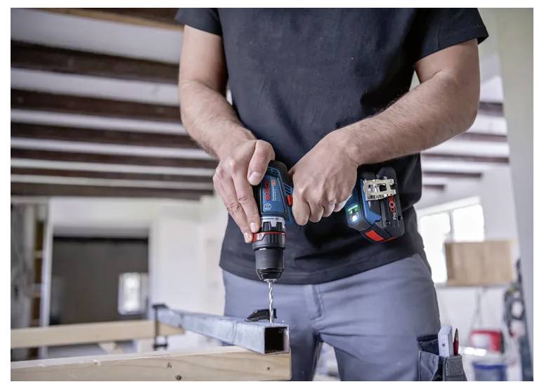 Eine Person verwendet eine kabellose Bohrmaschine, um ein Loch in einem Metallträger zu bohren und damit eine Bau- oder Heimwerkeraufgabe in einer Werkstattumgebung zu veranschaulichen.