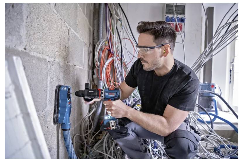 Eine Person mit Sicherheitsschutzbrille verwendet ein Elektrowerkzeug an einer Betonwand, umgeben von elektrischen Kabeln und Schaltschränken, was Bauarbeiten andeutet.