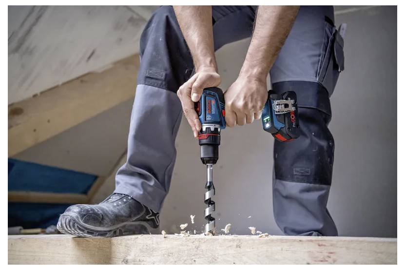 Eine Person, die mit einem kabellosen Bohrer ein Loch in Holz bohrt, trägt Arbeitskleidung und Sicherheitsstiefel, während Sägespäne auf der Oberfläche verstreut sind.