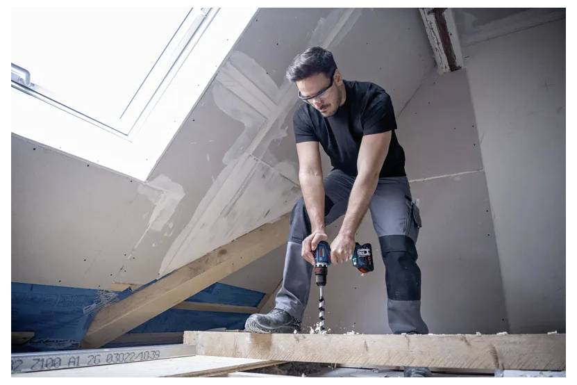 Eine Person, die einen Akkuschrauber verwendet, um eine Holzplanke in einem teilweise fertiggestellten Dachboden zu befestigen, trägt Schutzbrille und Arbeitskleidung.