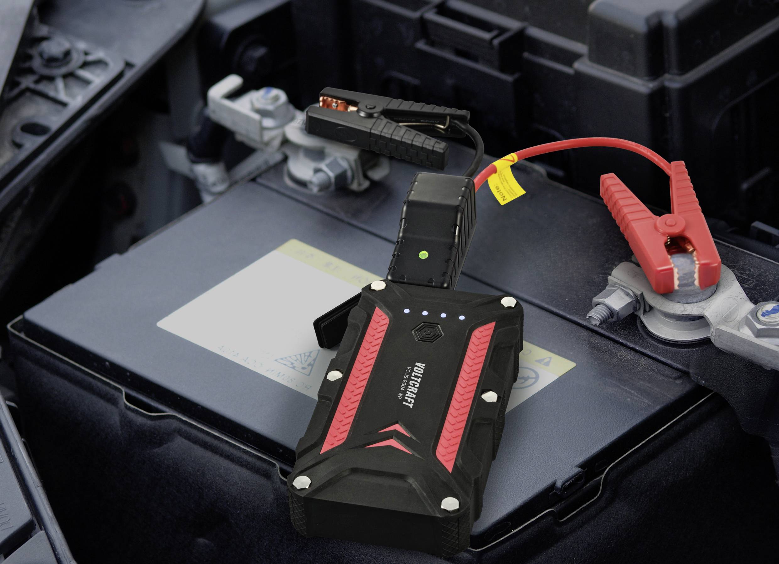 Sprungstarter und Überbrückungskabel sind an eine Autobatterie angeschlossen. Das Gerät zeigt eine grüne Anzeigeleuchte.