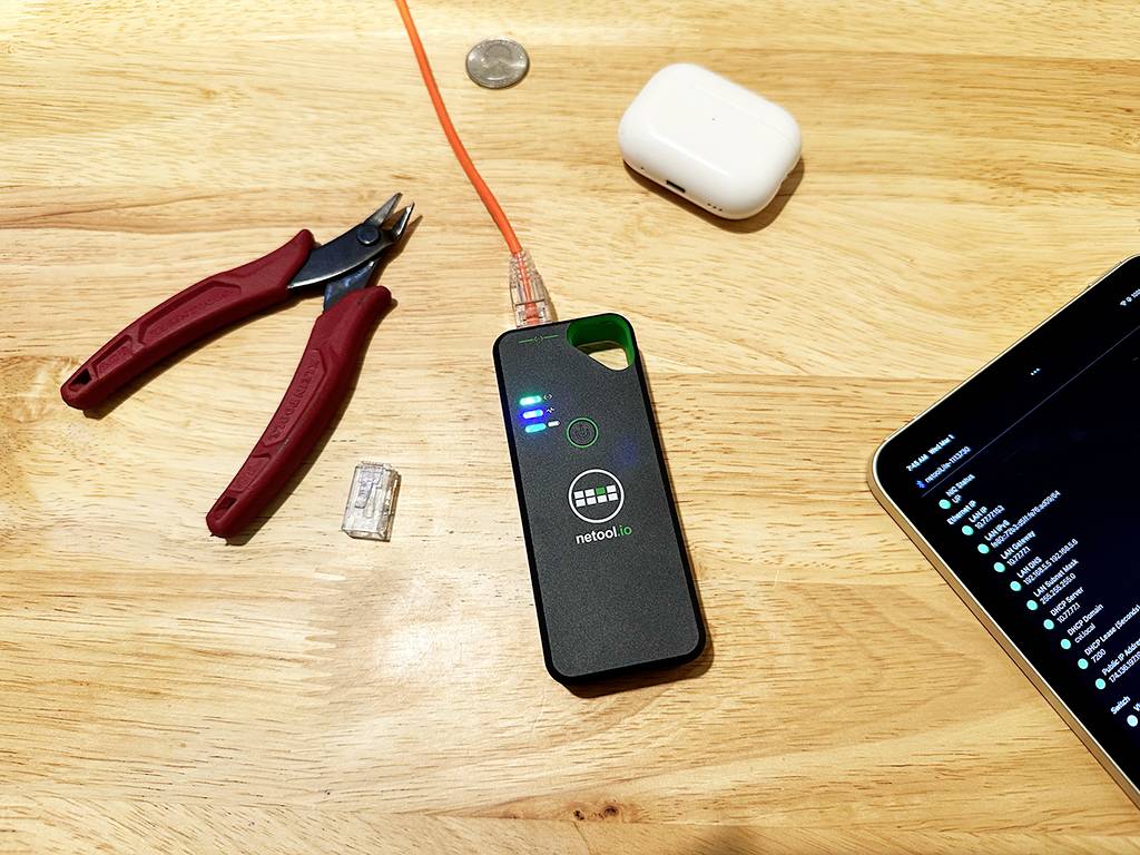 Auf einem Holztisch liegen ein Netzwerktester mit einer eingesteckten orangenen Kabel, daneben AirPods, eine Zange und ein Tablet.