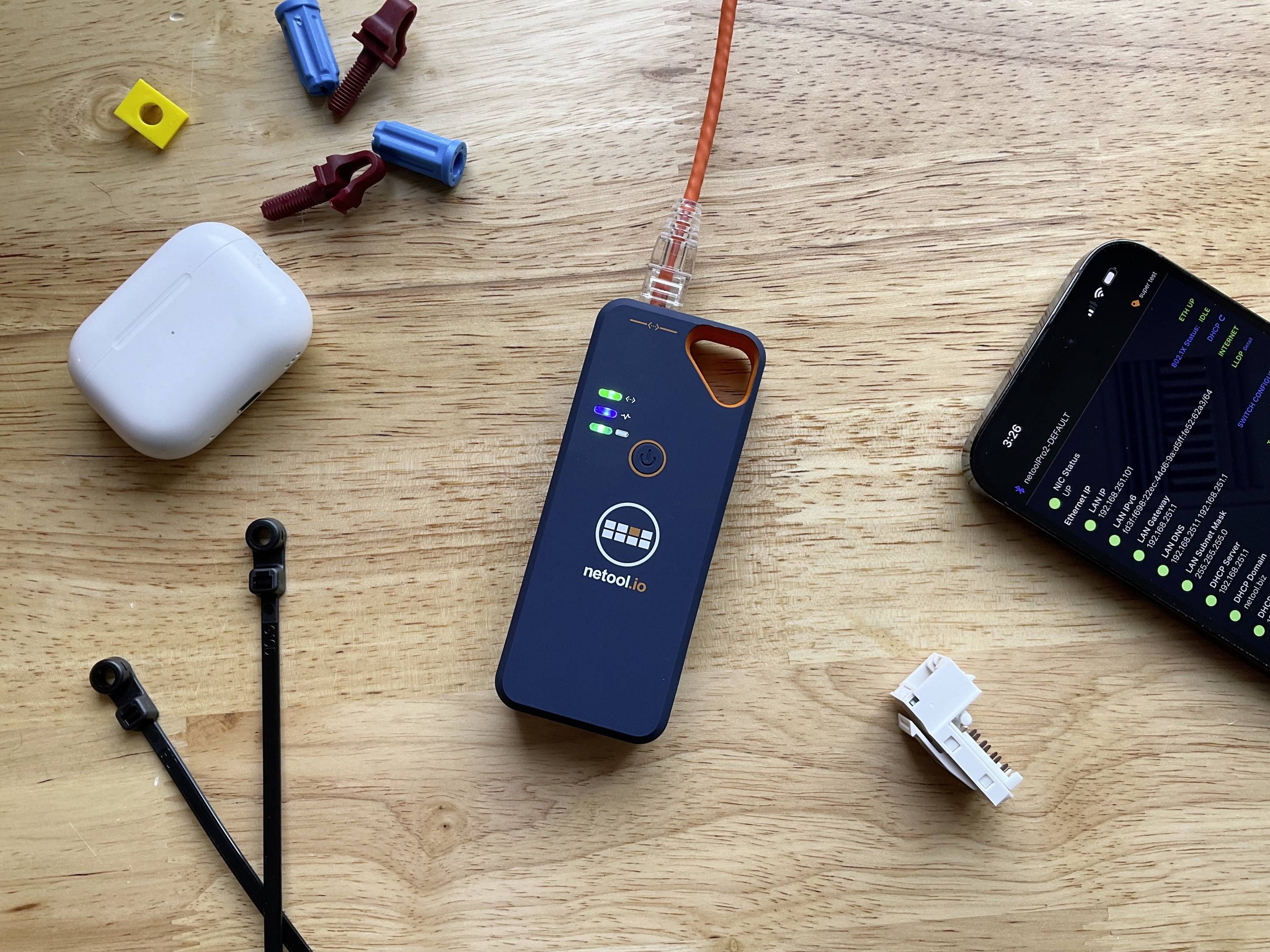 Ein Netzwerktestgerät mit leuchtenden LEDs liegt auf einem Tisch neben einem Smartphone, Kabelbindern und diversen Steckverbindern.