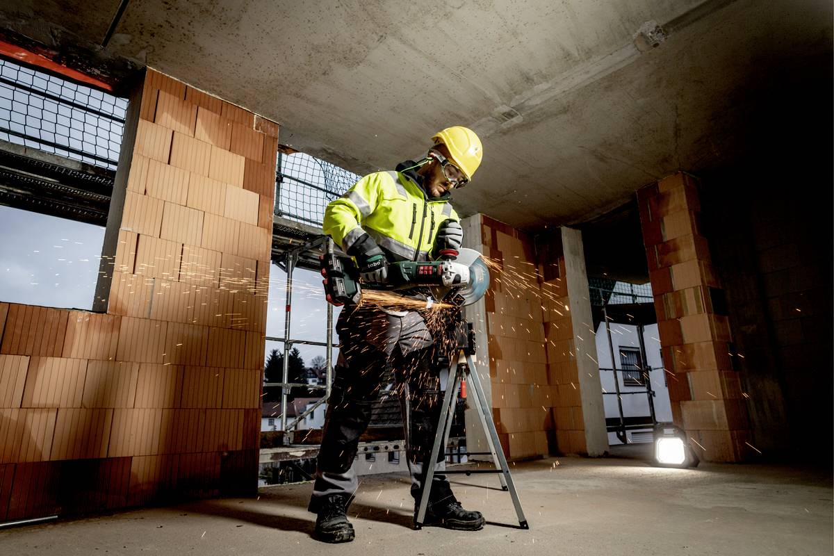 Ein Bauarbeiter in Schutzausrüstung schneidet Metall mit einem Winkelschleifer. Funken fliegen auf einer Baustelle.