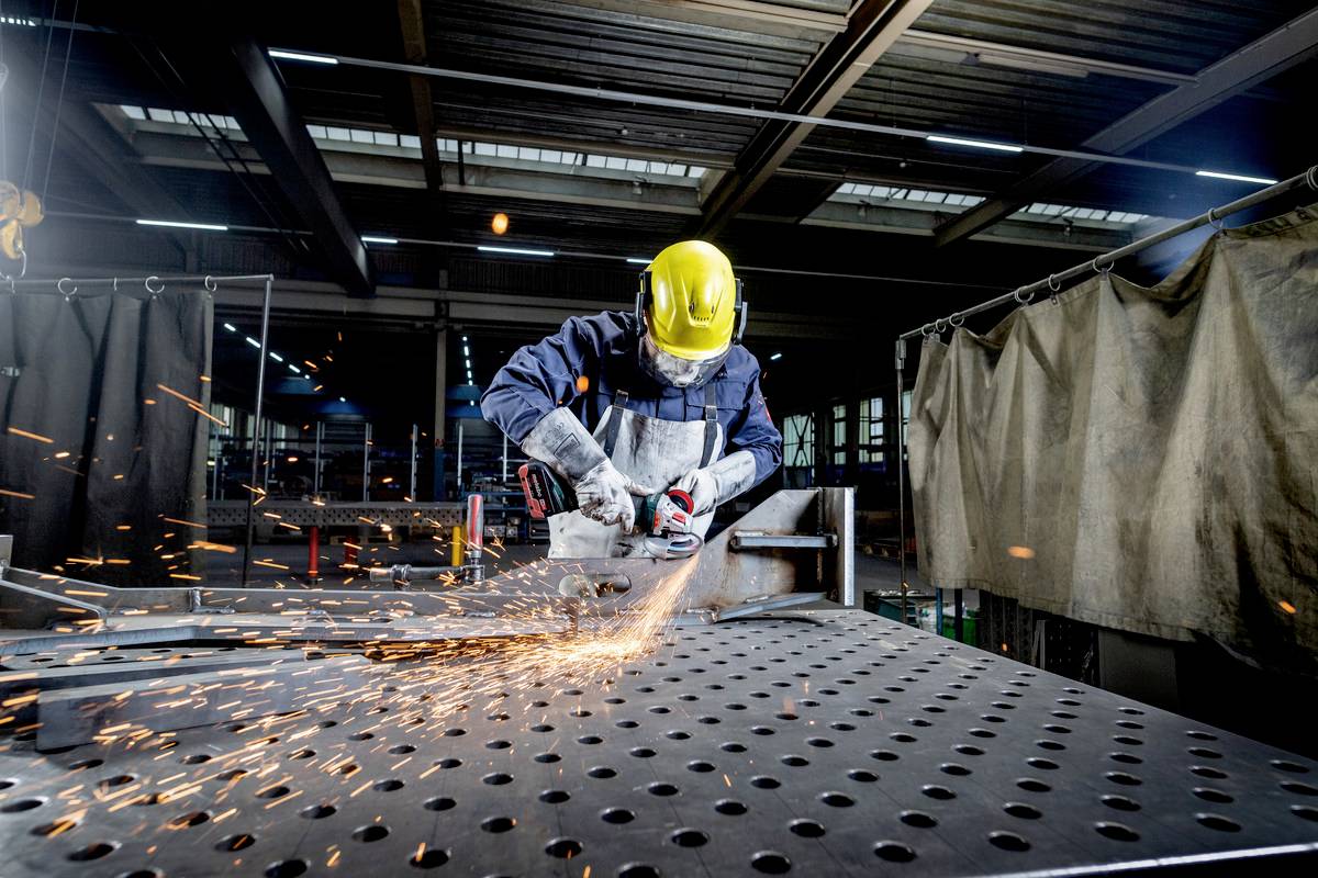 Ein Arbeiter mit gelbem Schutzhelm und Schutzbrille schleift Metall mit einem Winkelschleifer. Funken fliegen in einer Werkhalle.