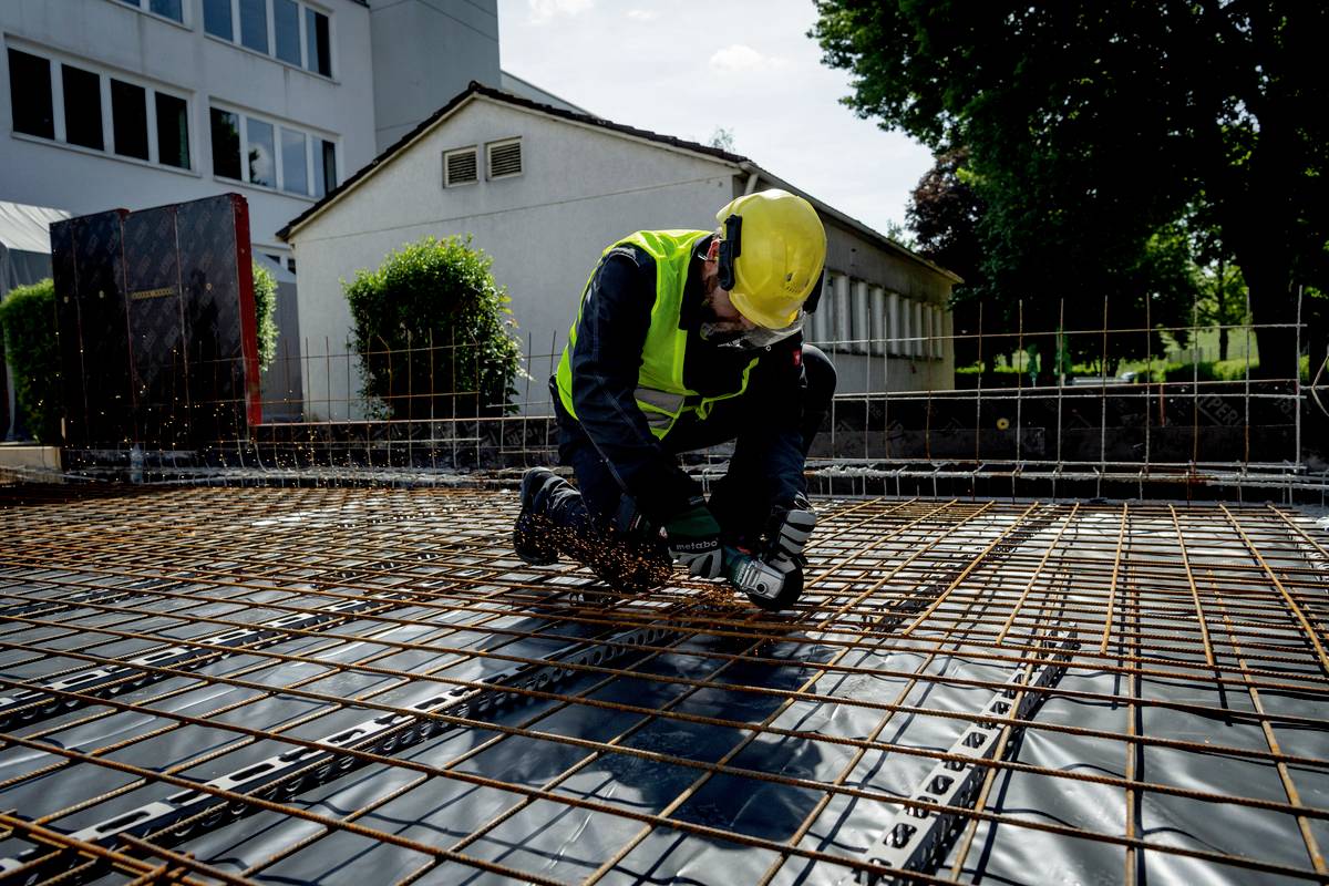 Bauarbeiter in Schutzkleidung schneidet Metallgitter auf einer Baustelle, umgeben von Gebäuden und Bäumen, bei Tageslicht.