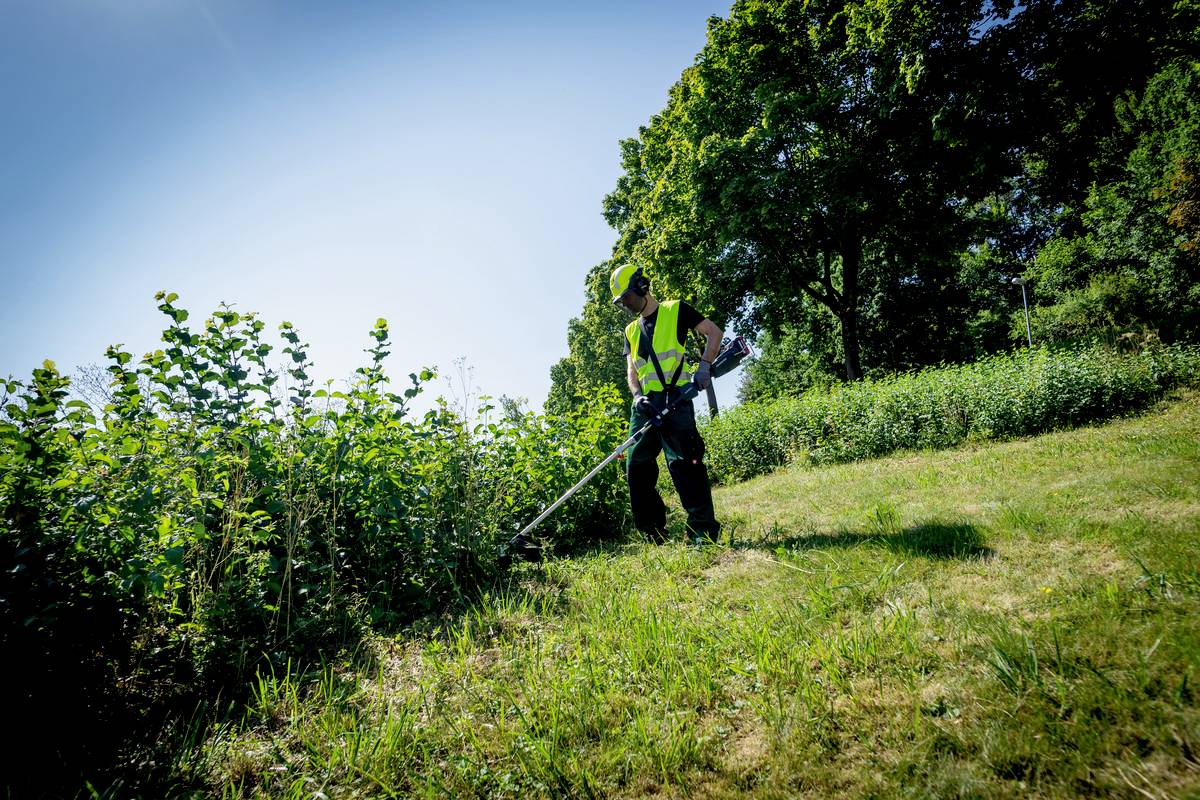 Eine Person in Warnweste schneidet mit einem Trimmer Gras auf einer Wiese. Im Hintergrund sind Bäume und blauer Himmel zu sehen.