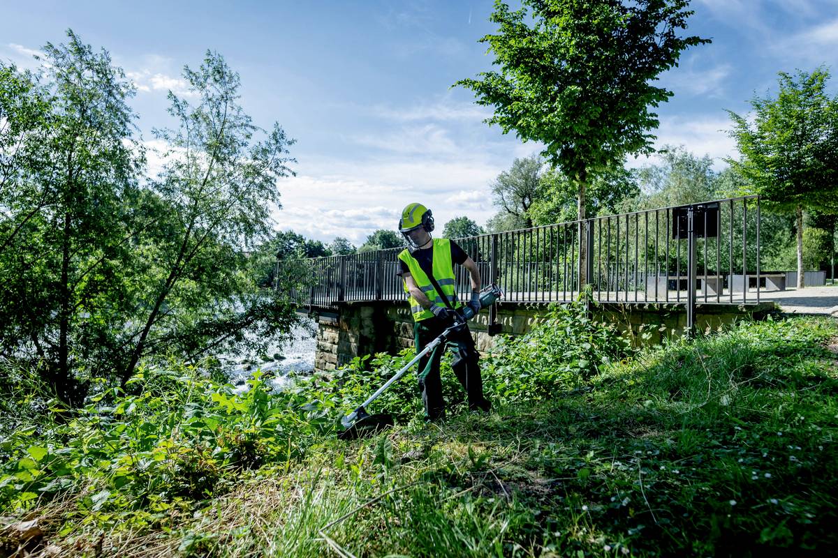 Eine Person in Arbeitskleidung schneidet mit einer Motorsense Gras und Unkraut neben einem Fluss, im Hintergrund eine Brücke.