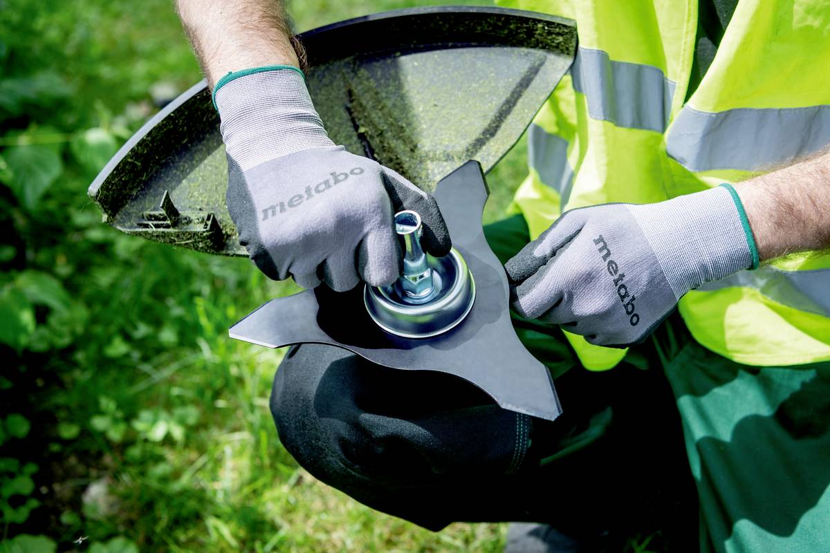 Eine Person trägt eine Sicherheitsweste und Handschuhe, repariert oder wechselt ein Schneidwerkzeug an einem Gartengerät im Freien.