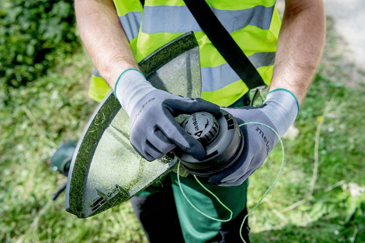 Eine Person trägt Handschuhe und hält einen Trimmer, bereit zum Einsatz in einem Garten. Gras und grüne Pflanzen im Hintergrund sichtbar.