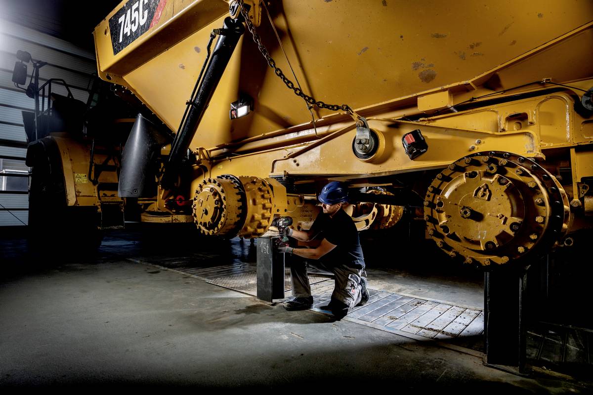 Ein Mechaniker arbeitet in einer Werkstatt mit einem großen gelben Baufahrzeug. Er überprüft Teile unter dem Fahrzeug.