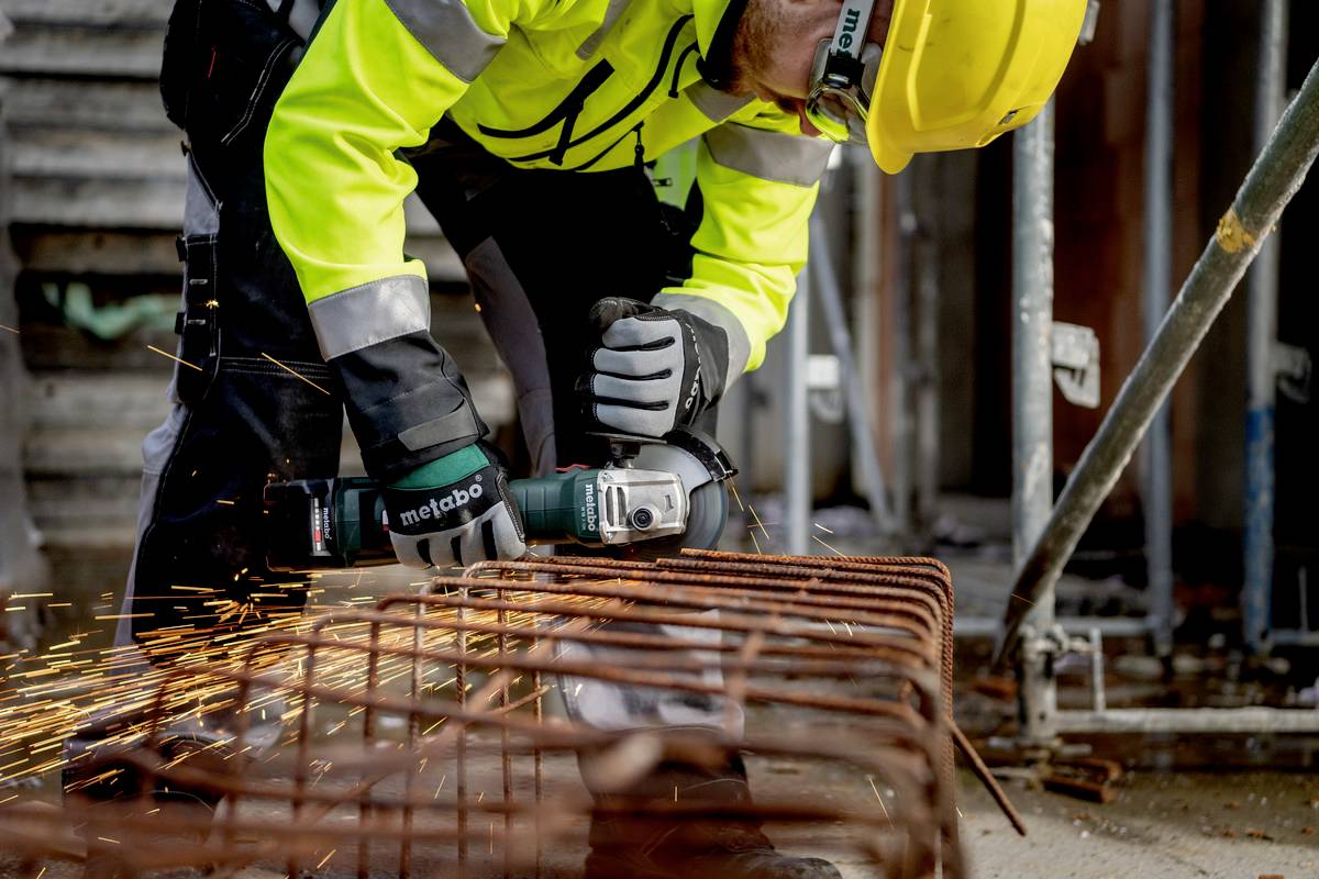 Eine Person mit gelbem Helm und Warnweste schneidet Metallstäbe mit einem Winkelschleifer, Funken fliegen im Arbeitsbereich.