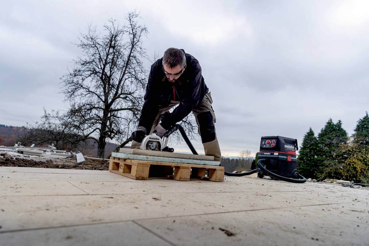 Person schneidet Holz auf einer Baustelle im Freien mit einer elektrischen Säge. Im Hintergrund sind Bäume und ein bewölkter Himmel.