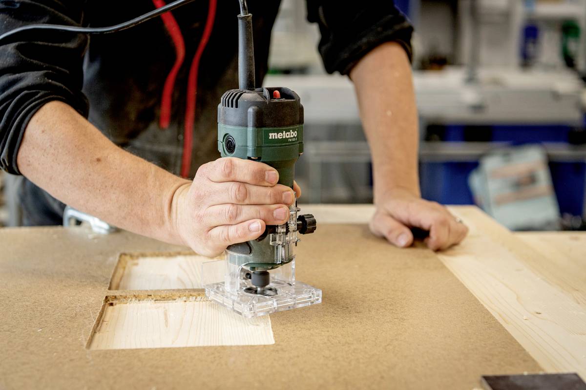 Person fräst ein rechteckiges Loch in eine Holzplatte mit einer Fräsmaschine in einer Werkstatt.