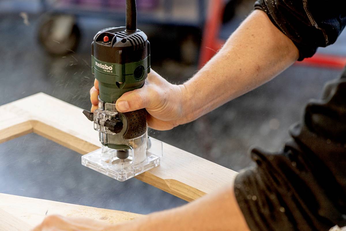 Eine Person verwendet eine Oberfräse, um eine Holzplatte zu bearbeiten. Holzspäne fliegen in der Werkstatt.