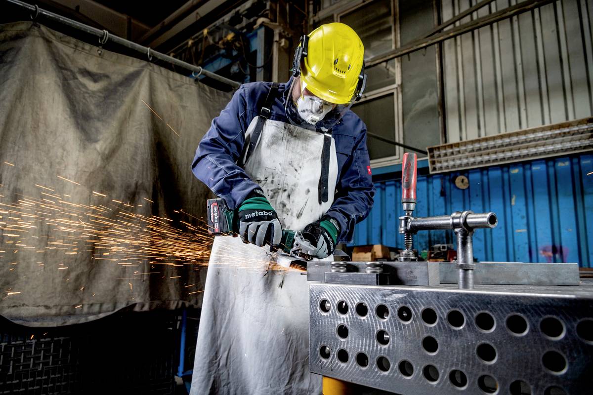 Ein Arbeiter in Schutzkleidung und gelbem Helm schleift Metall in einer Werkstatt. Funken fliegen vom Schleifgerät, umgeben von Maschinen.