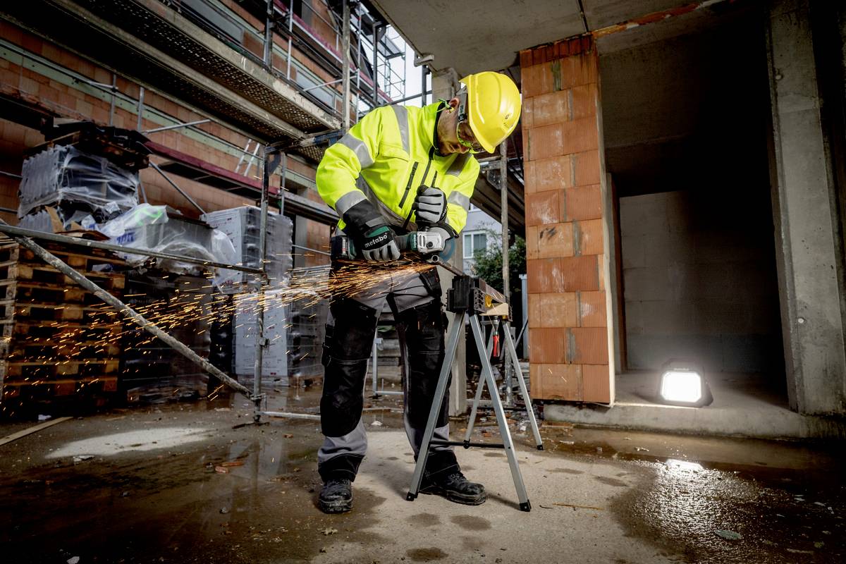 Ein Bauarbeiter mit Sicherheitskleidung schneidet Metall mit einer elektrischen Säge. Funken fliegen, Umgebung ist eine Baustelle.