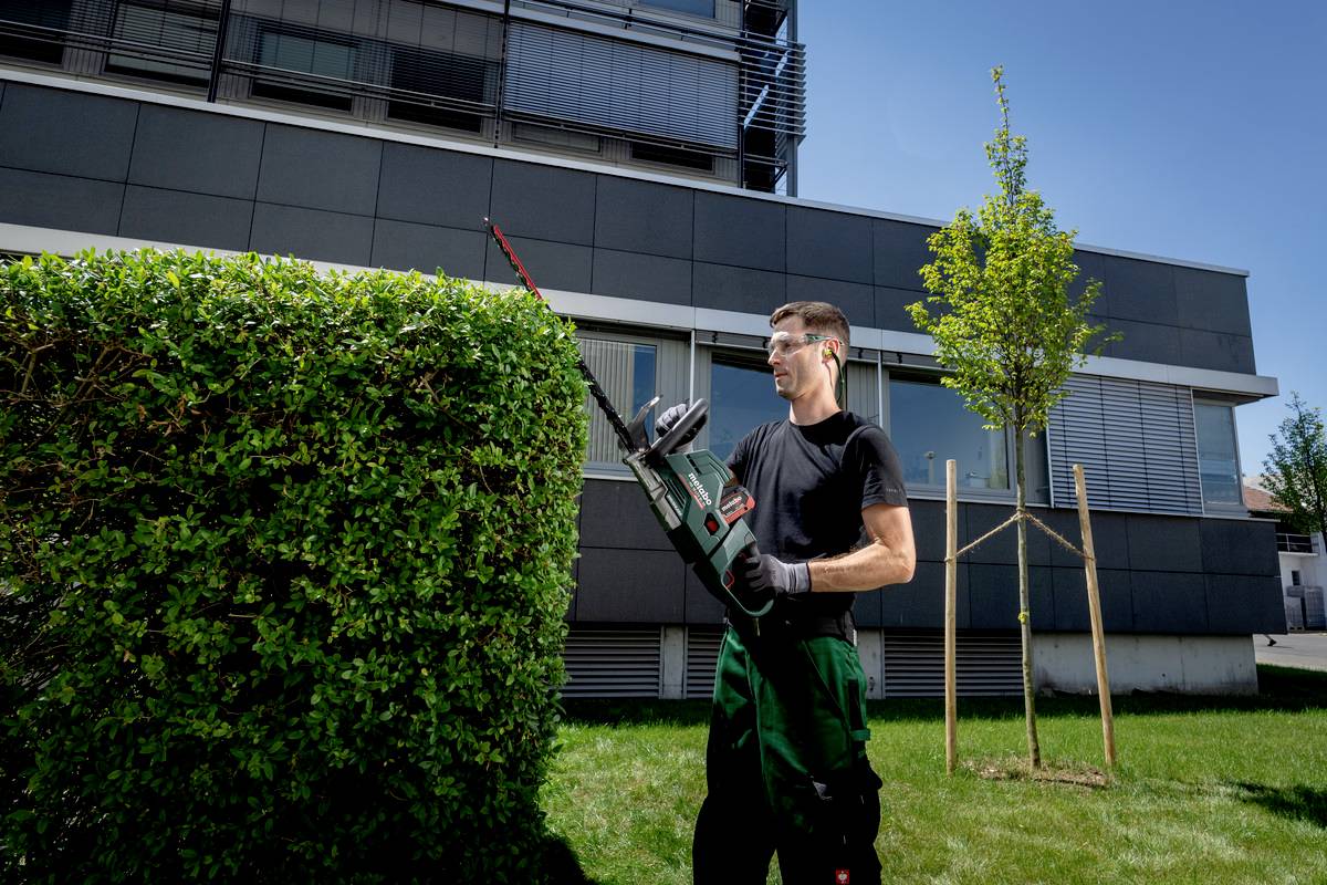 Ein Mann schneidet mit einer elektrischen Heckenschere eine Hecke vor einem modernen Gebäude. Er trägt Schutzhandschuhe und -brille.