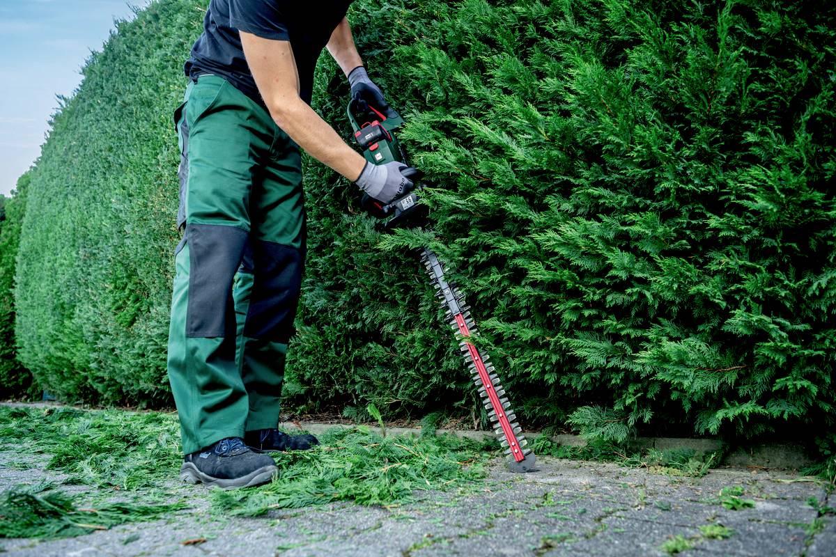 Eine Person schneidet eine Hecke mit einer elektrischen Heckenschere auf einem Gehweg.chnittgut liegt auf dem Boden.