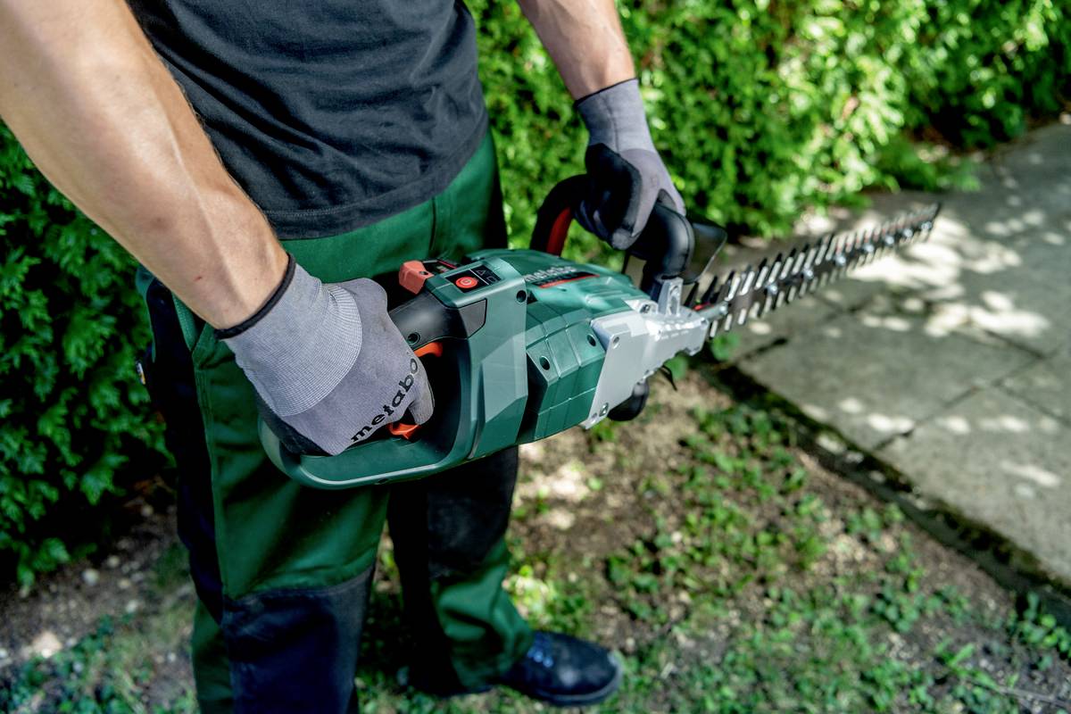 Person in Handschuhen hält eine elektrische Heckenschere und beschneidet eine Hecke im Garten.