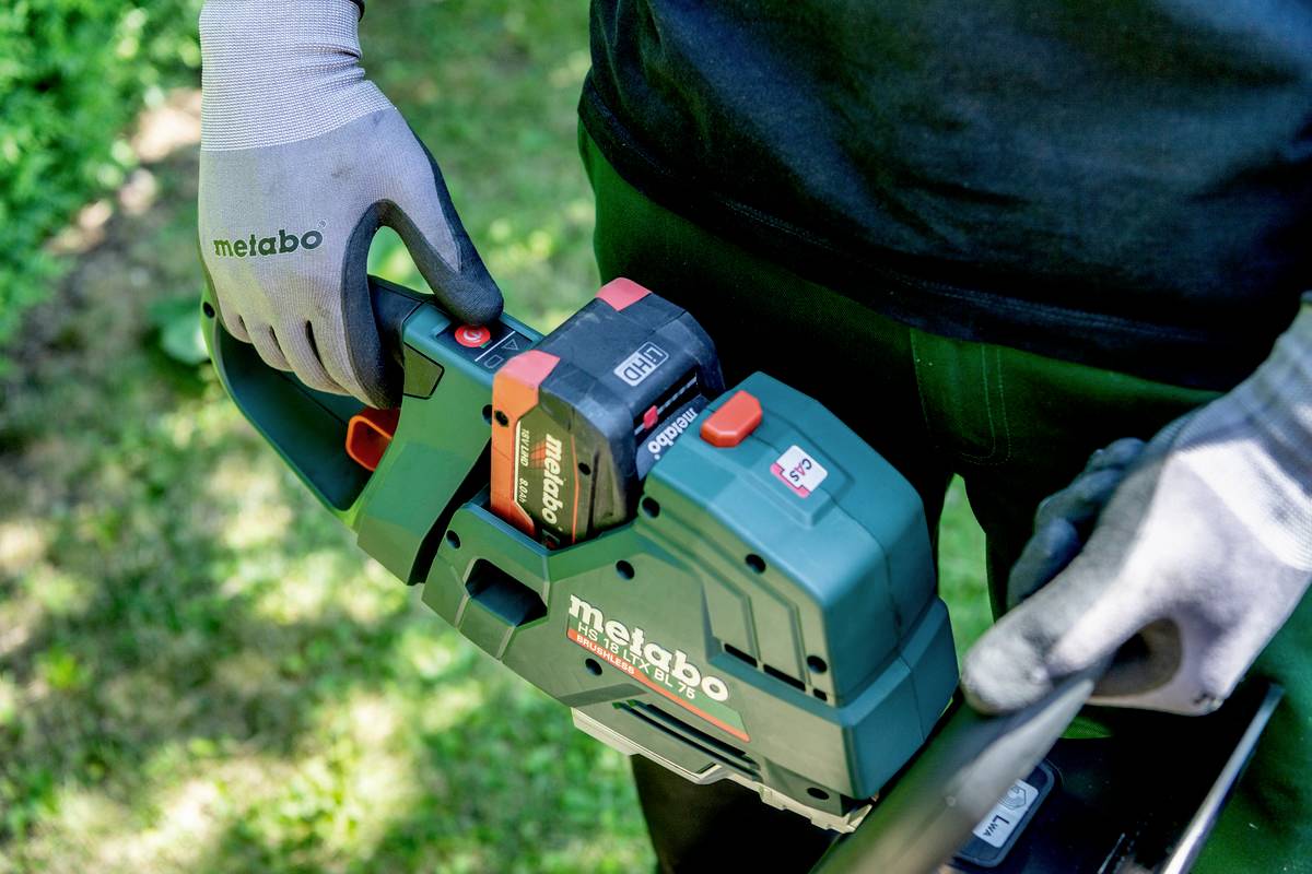 Person mit Arbeitshandschuhen hält eine grüne Elektrosäge der Marke Metabo, die im Garten verwendet wird.