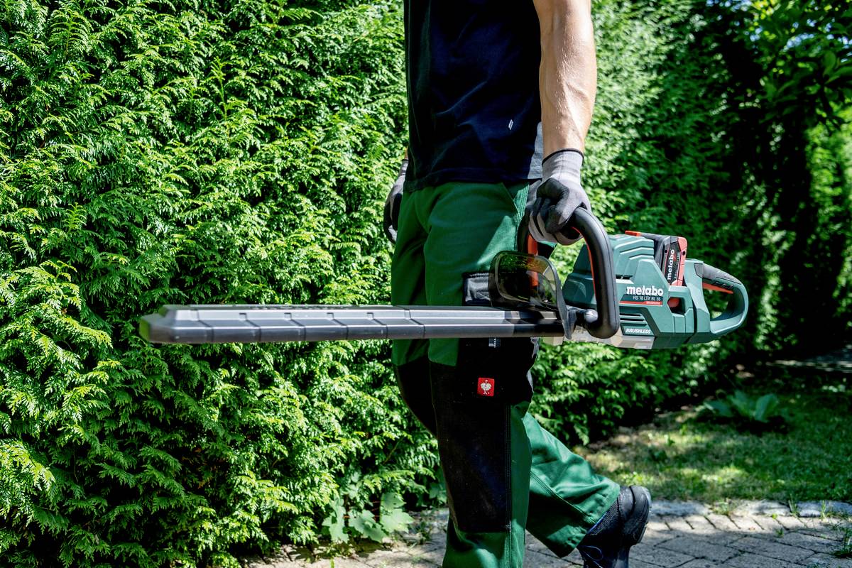 Eine Person in Arbeitskleidung hält eine elektrische Heckenschere neben einer gepflegten, grünen Hecke.