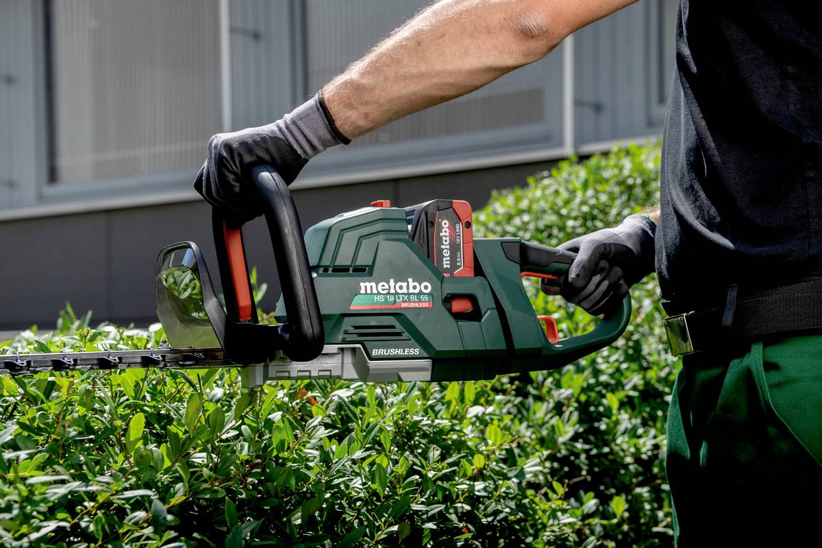 Eine Person schneidet mit einer Metabo-Heckenschere eine Hecke. Die Heckenschere ist grün und schwarz, mit roten Elementen.