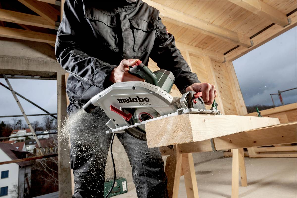 Person sägt ein Holzbrett mit einer elektrischen Handkreissäge in einer Baustelle. Draußen ist bewölktes Wetter sichtbar.