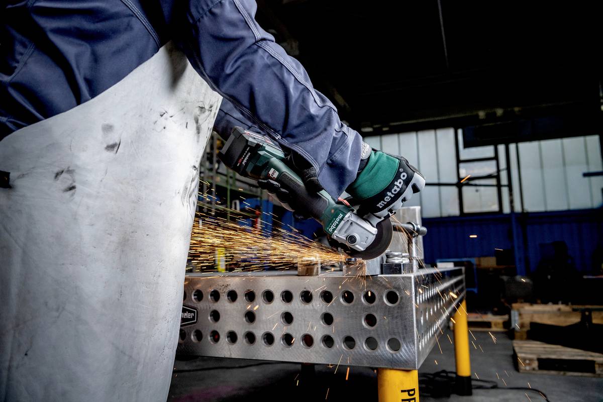Eine Person schleift Metall mit einem Winkelschleifer in einer Werkstatt, Funken fliegen. Schutzkleidung und Handschuhe werden getragen.
