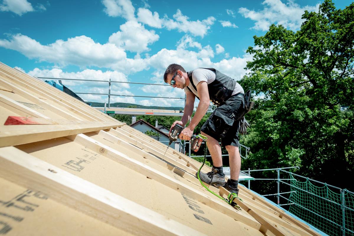 Ein Bauarbeiter arbeitet auf einem schrägen Dach unter blauem Himmel; er verwendet Werkzeuge und trägt Sicherheitsausrüstung.