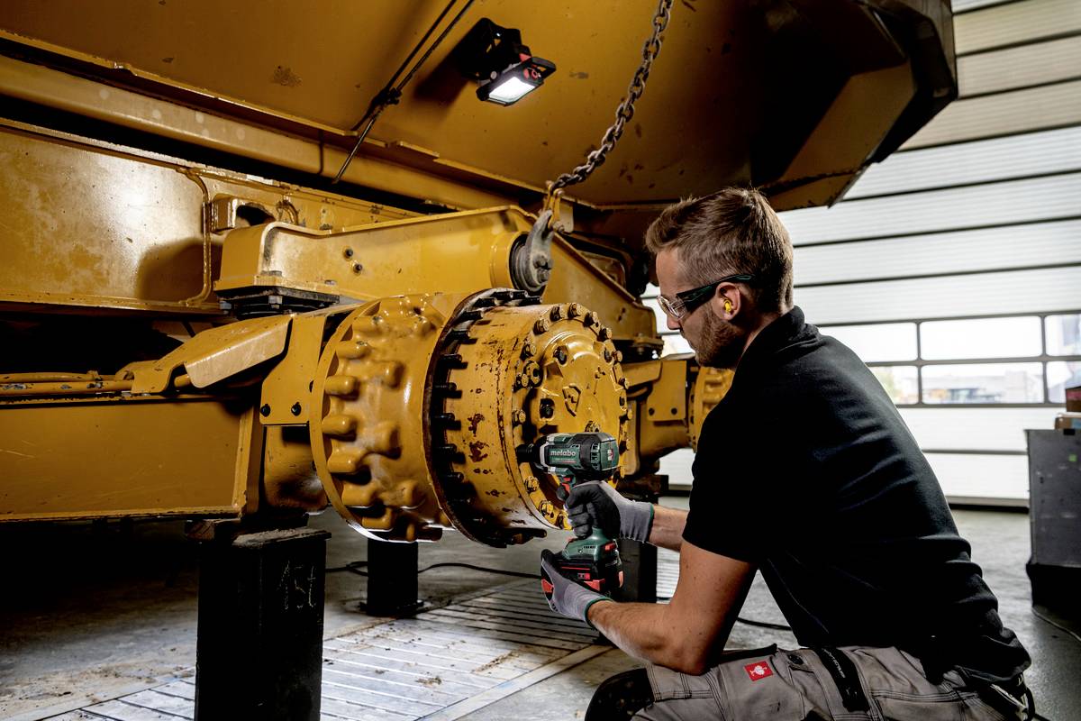 Ein Mann in Arbeitskleidung verwendet ein Elektrowerkzeug, um an einem großen gelben Maschinenteil zu arbeiten, in einer Werkstatt-Umgebung.