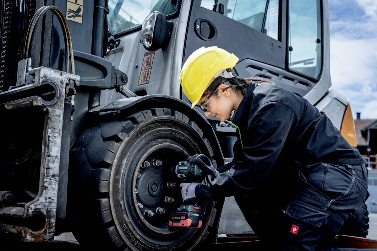 Eine Person in Arbeitskleidung und Schutzhelm repariert das Rad eines schweren Fahrzeugs mit einem Elektrowerkzeug.