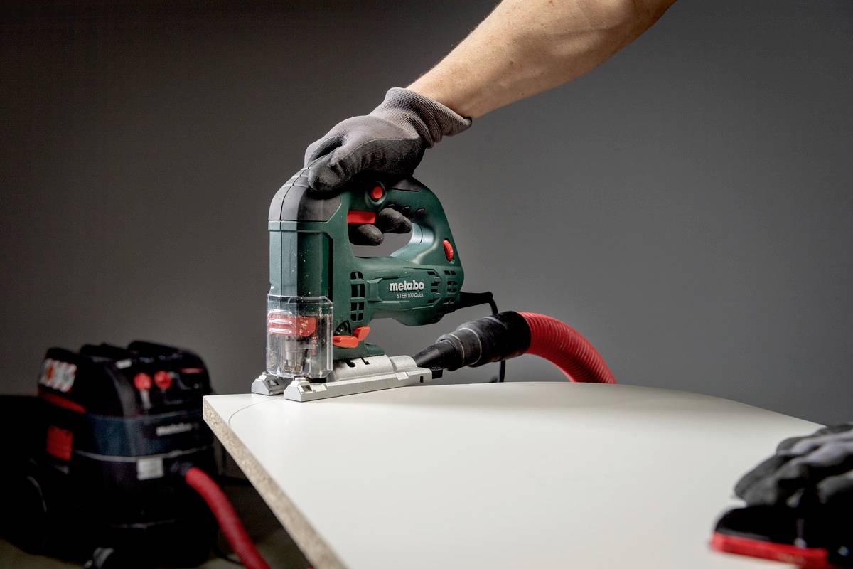 Eine Person schneidet ein Stück Holz mit einer grünen Stichsäge. Ein Industriesauger ist angeschlossen, um Staub abzuleiten.