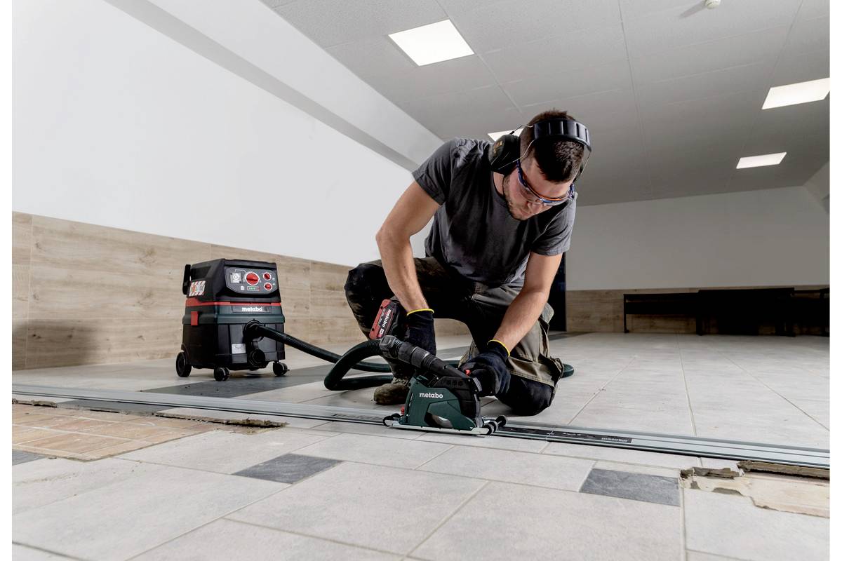 Eine Person trägt Gehörschutz und bedient eine Fliesenschneidemaschine in einem Raum mit hellen Wänden und gefliestem Boden.
