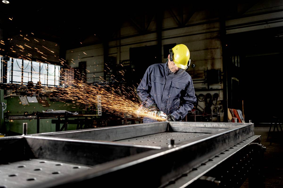 Ein Arbeiter in einer Werkstatt schleift Metall, während Funken sprühen. Er trägt einen gelben Helm und Gehörschutz.