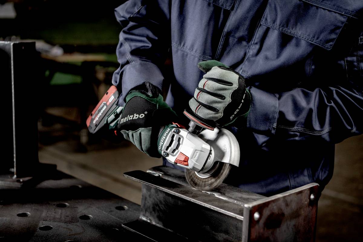 Ein Handwerker in Arbeitskleidung schleift Metall mit einem Winkelschleifer in einer Werkstatt. Hände sind in Schutzhandschuhen.