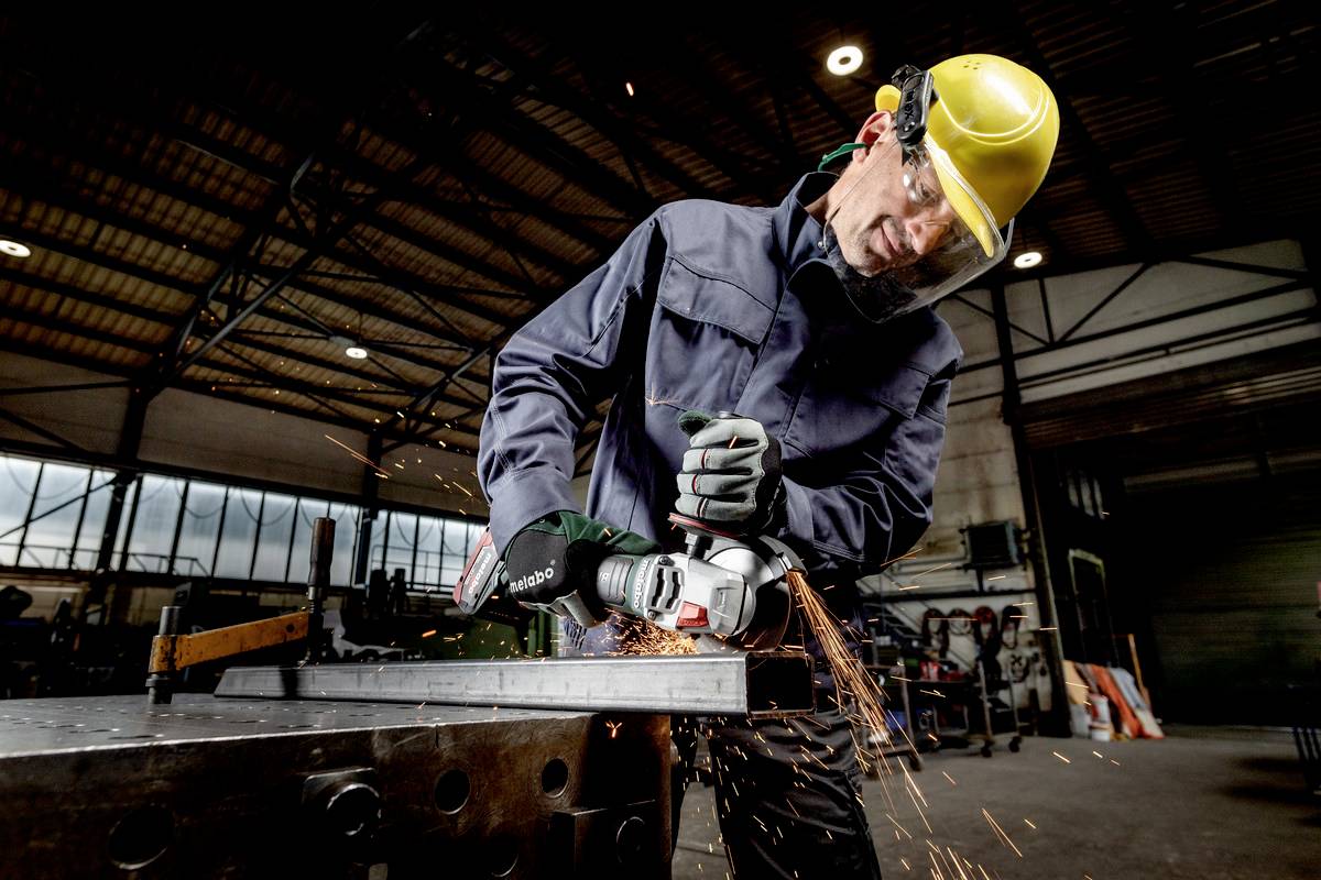 Arbeiter in Schutzkleidung schleift Metall mit einem Winkelschleifer in einer Werkhalle, Funken fliegen.