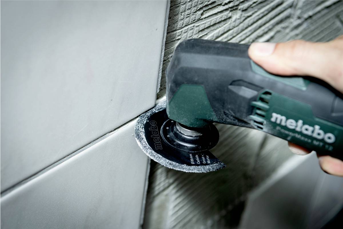 Hand hält elektrisches Werkzeug mit kleiner Säge, schneidet Fugen zwischen Wandfliesen. Gesicht der Fliesen und Fugen sichtbar.